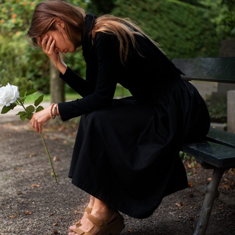 Frau in schwarzem Kleid sitzt auf einer Bank, hält eine Blume und zeigt Traurigkeit.